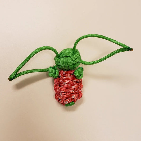 A small decorative knot made from red and green paracord, resembling a strawberry with green leaves. The green cord forms a double loop at the top, resembling the knot’s stem and leaves, while the red cord is intricately woven to represent the body of the strawberry—a charming example of Green Alien Baby - Handmade Paracord Alien Doll - Mint & Strawberry - 550 Paracord - Made in USA by Insanely Paracord creations.