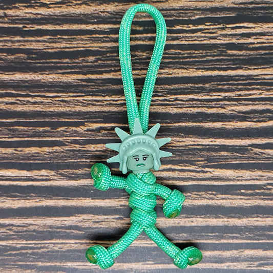 A durable NYC Statue of Liberty Paracord Pal Keychain 550 Cord Strength 2.5 Feet Mint, this small figurine made of green 550 paracord features a face and crown resembling the iconic statue. With arms, legs, and a loop for hanging, it rests on a dark, textured wooden surface.