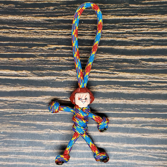 A colorful, twisted lanyard made from 550 Paracord with red, blue, and yellow strands forms a stick figure with a loop at the top. The figure's head is a small, angry face with short brown hair. The Insanely Paracord Scary Movie Serial Killer Keychain with 550 Paracord has a background featuring a wood grain texture.