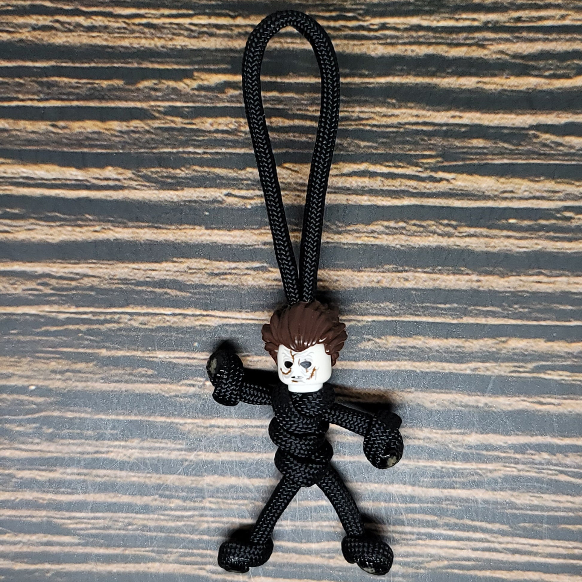 A small figurine with a rough, cartoonish face and spiky brown hair attached to a black paracord rope sits against a wooden surface. This Mike Scary Movie Serial Killer Halloween Paracord Pal Keychain by Insanely Paracord figure is tied to resemble a stick person, making it an eerie and playful accessory for horror movie fans.