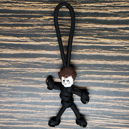 A small figurine with a rough, cartoonish face and spiky brown hair attached to a black paracord rope sits against a wooden surface. This Mike Scary Movie Serial Killer Halloween Paracord Pal Keychain by Insanely Paracord figure is tied to resemble a stick person, making it an eerie and playful accessory for horror movie fans.