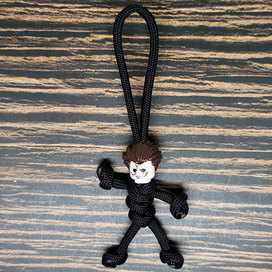 A small figurine with a rough, cartoonish face and spiky brown hair attached to a black paracord rope sits against a wooden surface. This Mike Scary Movie Serial Killer Halloween Paracord Pal Keychain by Insanely Paracord figure is tied to resemble a stick person, making it an eerie and playful accessory for horror movie fans.