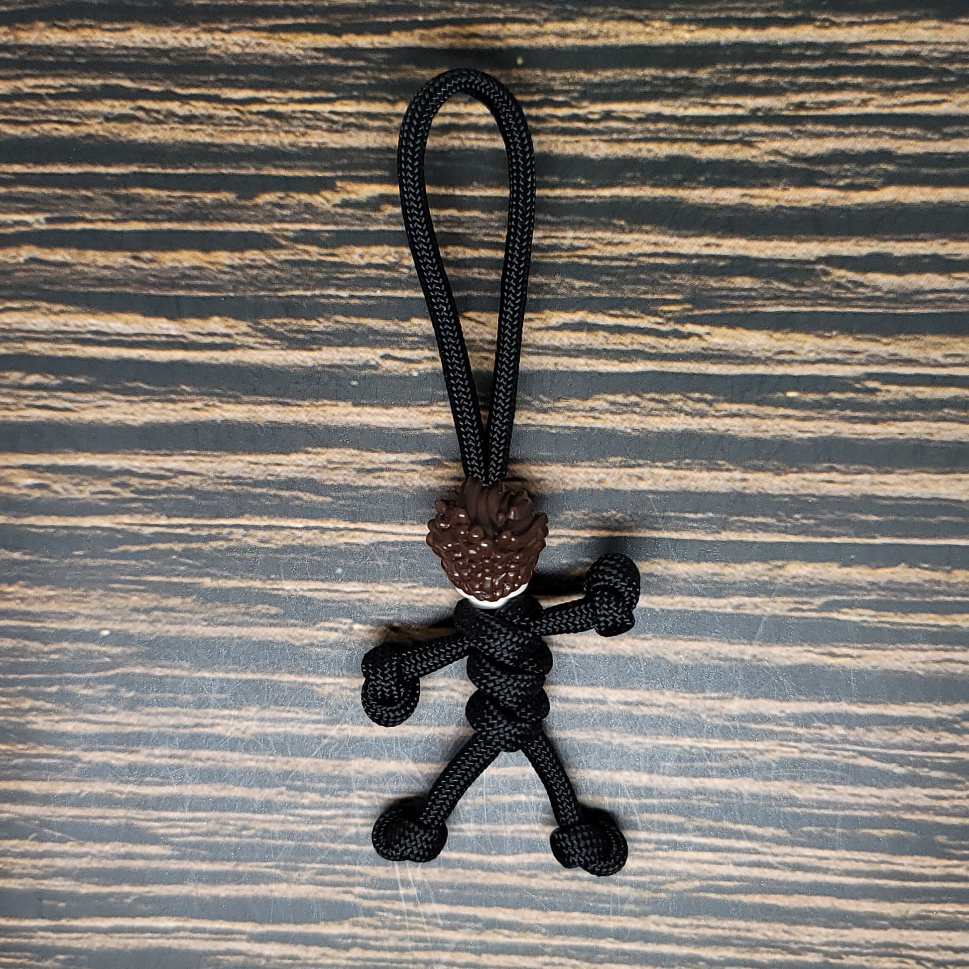 A small, handmade figurine crafted from black paracord, featuring a loop at the top for hanging. The figure has detailed hair made from brown paracord and is posed as though running or in motion, set against a textured, striped background—ideal as an Insanely Paracord Mike Scary Movie Serial Killer Halloween Paracord Pal Keychain.