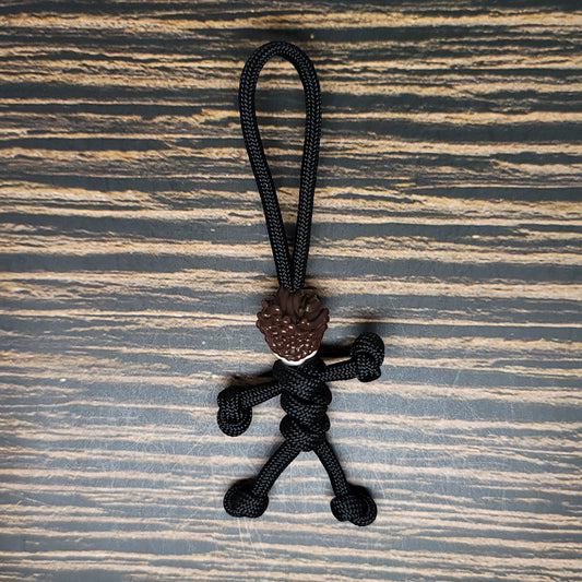 A small, handmade figurine crafted from black paracord, featuring a loop at the top for hanging. The figure has detailed hair made from brown paracord and is posed as though running or in motion, set against a textured, striped background—ideal as an Insanely Paracord Mike Scary Movie Serial Killer Halloween Paracord Pal Keychain.