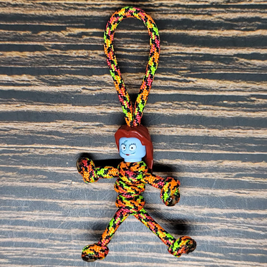 A small handmade Halloween accessory made with colorful twisted rope features a blue plastic head with red hair. The Nightmare Pumpkin Queen Keychain | Handmade Paracord Pal by Insanely Paracord has a loop at the top for hanging and is displayed on a textured wooden surface.