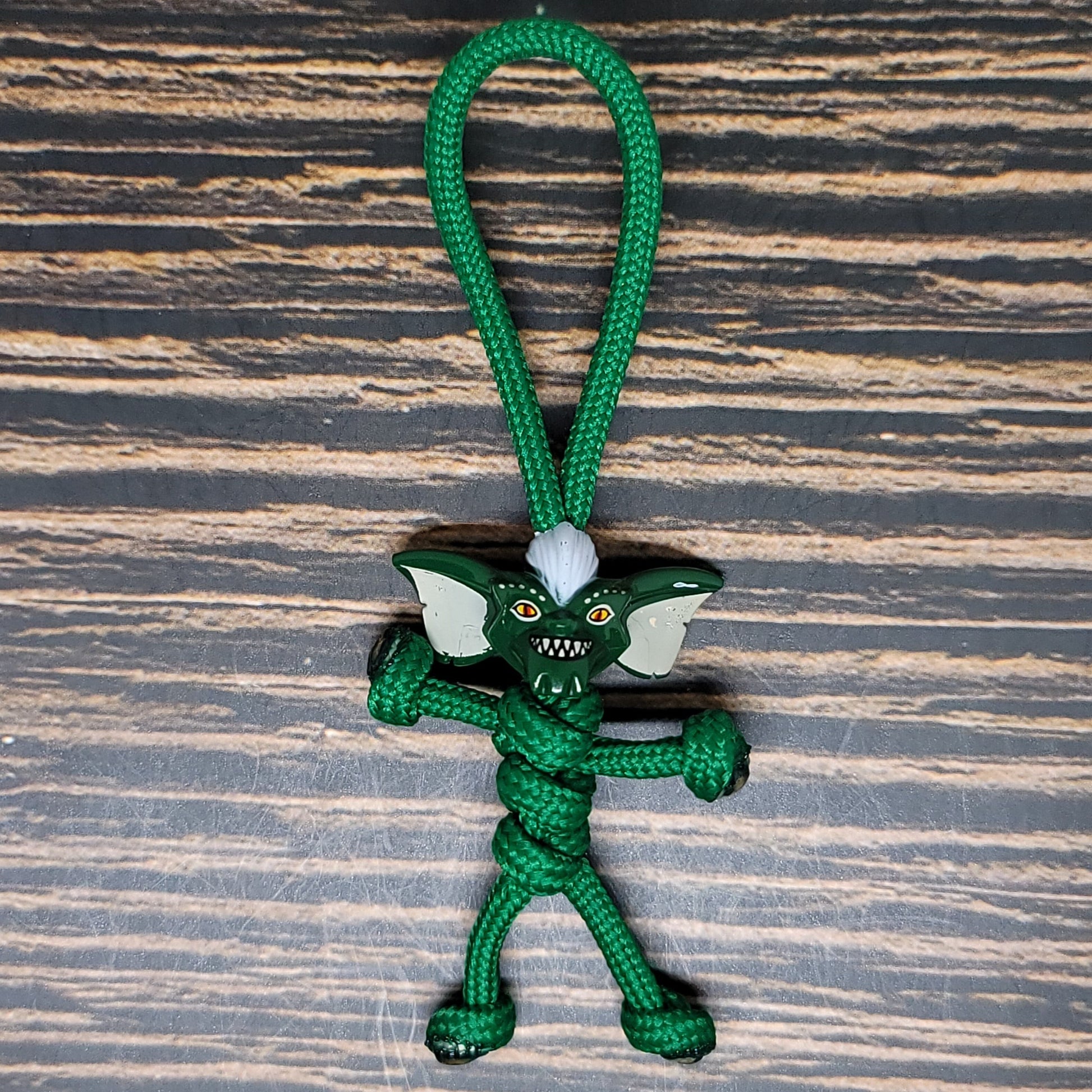 A small figurine made with green paracord, resembling a gremlin with large ears, sharp teeth, and a mischievous expression, lies on a wooden surface. This handcrafted Paracord Pal Keychain - 550 Paracord, Handmade in USA, Dark Green, High Strength by Insanely Paracord is intricately knotted to form the body, limbs, and head of the character.