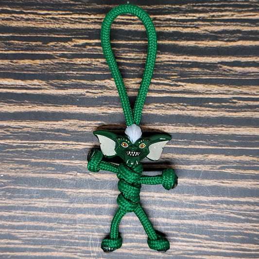 A small figurine made with green paracord, resembling a gremlin with large ears, sharp teeth, and a mischievous expression, lies on a wooden surface. This handcrafted Paracord Pal Keychain - 550 Paracord, Handmade in USA, Dark Green, High Strength by Insanely Paracord is intricately knotted to form the body, limbs, and head of the character.