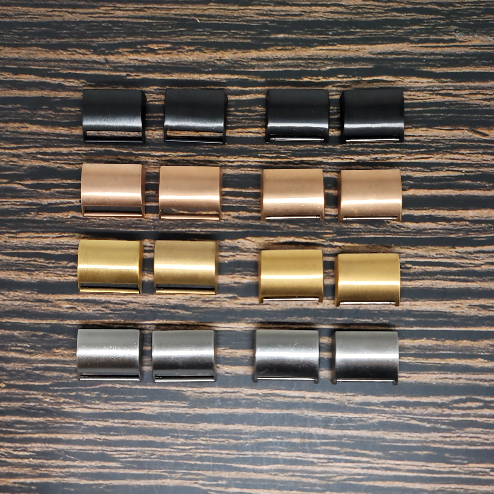 A collection of Insanely Paracord Fitbit Watch Band Adapter - Convert Your Smartwatch with Adaptors in Four Colors, compatible with Fitbit models, arranged in four rows on a wooden surface. Each row contains four adaptors of a different color: black, copper, gold, and silver.