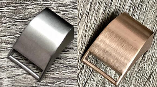 Two metal belt buckles are shown side by side on a textured wooden surface. The buckle on the left is matte silver, while the one on the right is a polished bronze color. Both have a sleek, curved design and an open rectangular slot at the bottom for attaching Insanely Paracord's Fitbit Alta Watch Band - Paracord, High-Quality, USA Made.